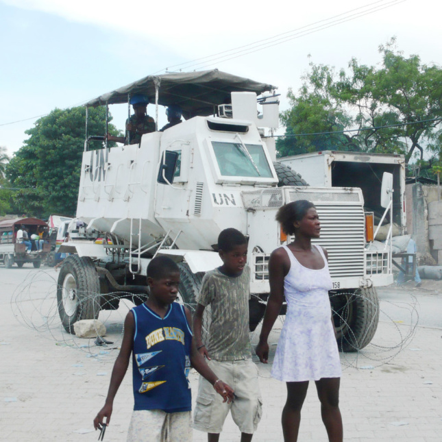 在海地太子港，人们站在部署在联合国-联海稳定团(UN-MINUSTAH)维和任务的装甲运兵车(APC)前。这辆装甲运兵车被带刺的铁丝网包围，并安装在轮毂上，至少暂时从一辆车辆转变为一种准建筑技术——移动塔。