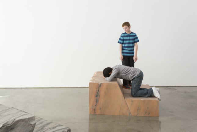 一个小男孩跪在大理石底座上，另一个男孩俯瞰着他的艺术装置照片