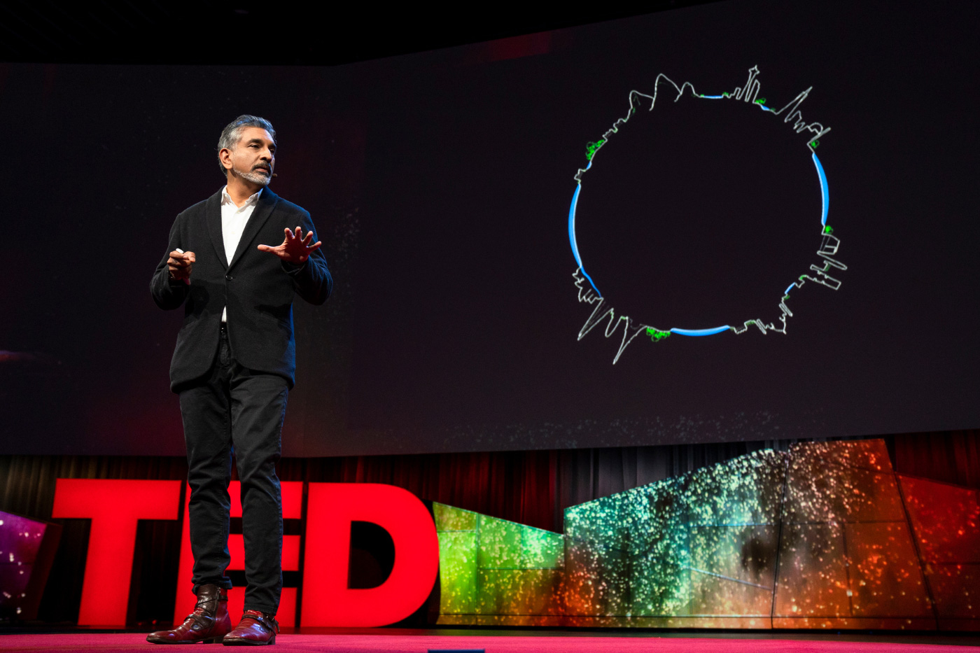 一个身穿黑色西装的印度人站在舞台上，上面用大字写着“TED”