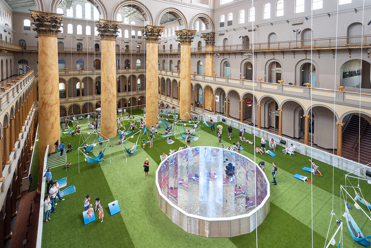 鸟瞰国家建筑博物馆的大厅，有柱子和绿色地毯，人们在蓝色吊床和草坪椅上闲逛
