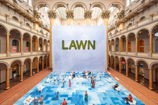 照片上的白色和蓝色的大招牌，上面印着“草坪”两个字，下面是蓝色格子地毯，人们在国家建筑博物馆闲逛