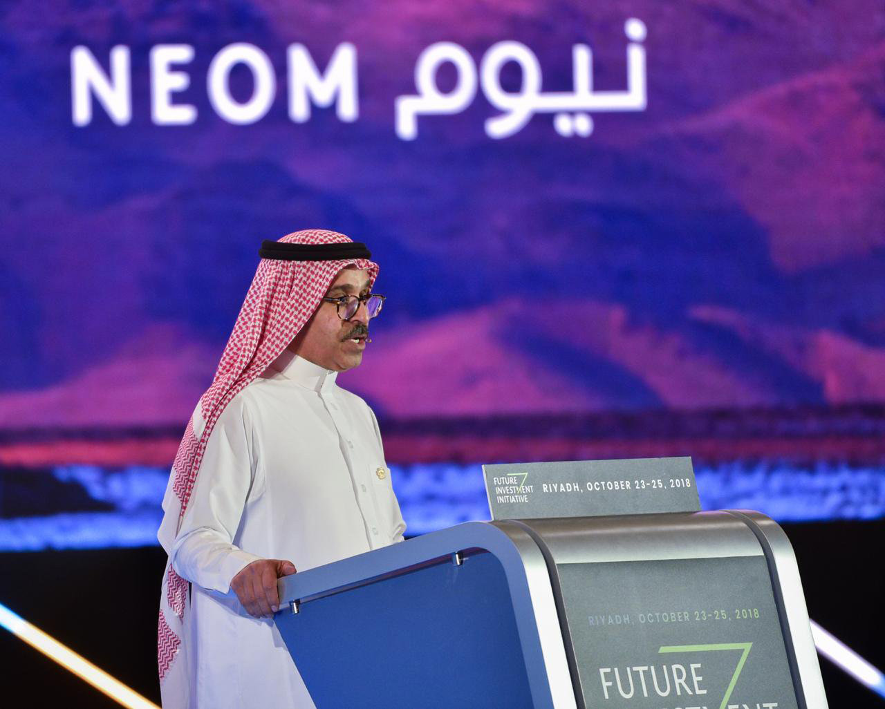 男子在讲台上讲话的图像，他身后有紫色的标志，用英语和阿拉伯语写着NEOM