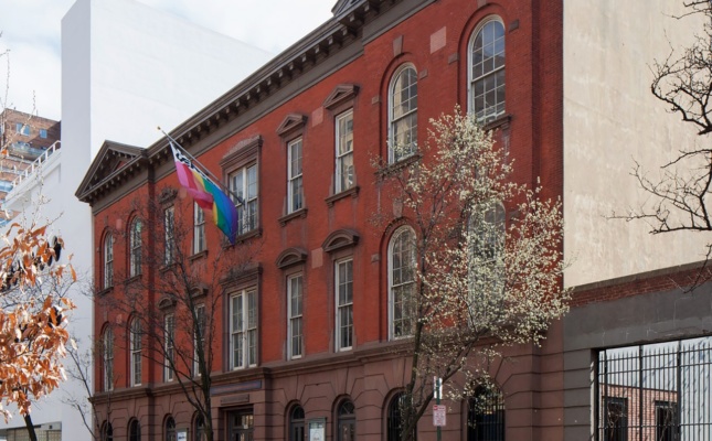在绿树成荫的街道上，一座砖砌建筑的透视照片。一面彩虹旗在大楼的入口处飘扬。