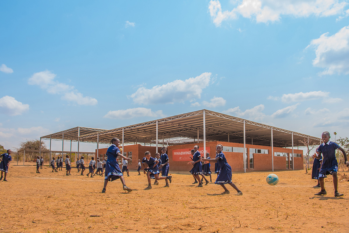 非洲儿童在砖砌学校前踢足球的画面，学校的招牌上写着“Mwabwindo”