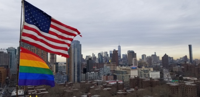 一面美国国旗和彩虹旗在布鲁克林一栋建筑的顶部飘扬，可以看到布鲁克林市中心和曼哈顿下城的景色