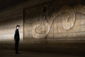 这是2021年沙丘电影的剧照，一个年轻人站在一个巨大的蠕虫神的金色浮雕前